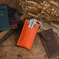 A set of personalized leather makeup brush pouches displayed on a rustic wooden table with dried flowers. The central, most prominent pouch is bright orange and contains several silver makeup brushes.