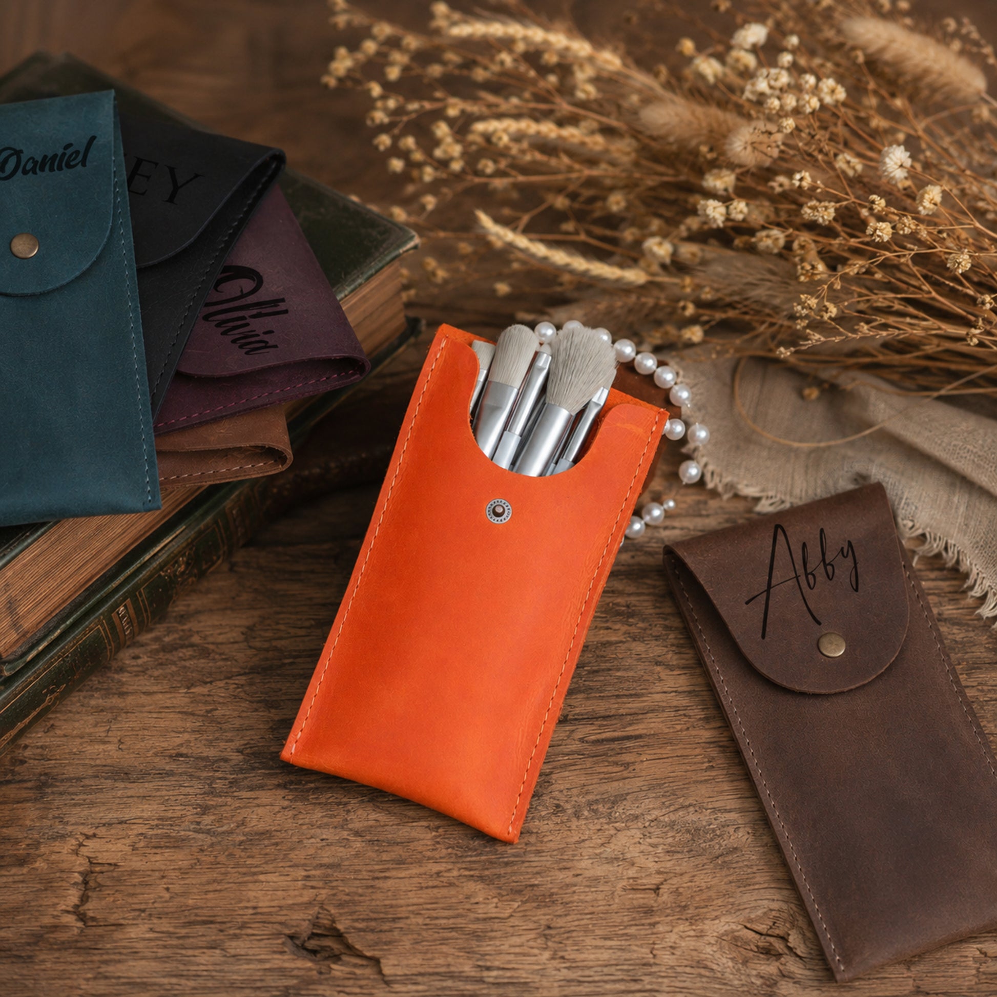 A set of personalized leather makeup brush pouches displayed on a rustic wooden table with dried flowers. The central, most prominent pouch is bright orange and contains several silver makeup brushes.