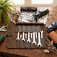 A brown leather tool roll laid open on a wooden table, holding several silver wrenches. The scene includes a potted fern, dried flowers, toy cars, a black vintage car drawing, and metal wrenches as styled props.