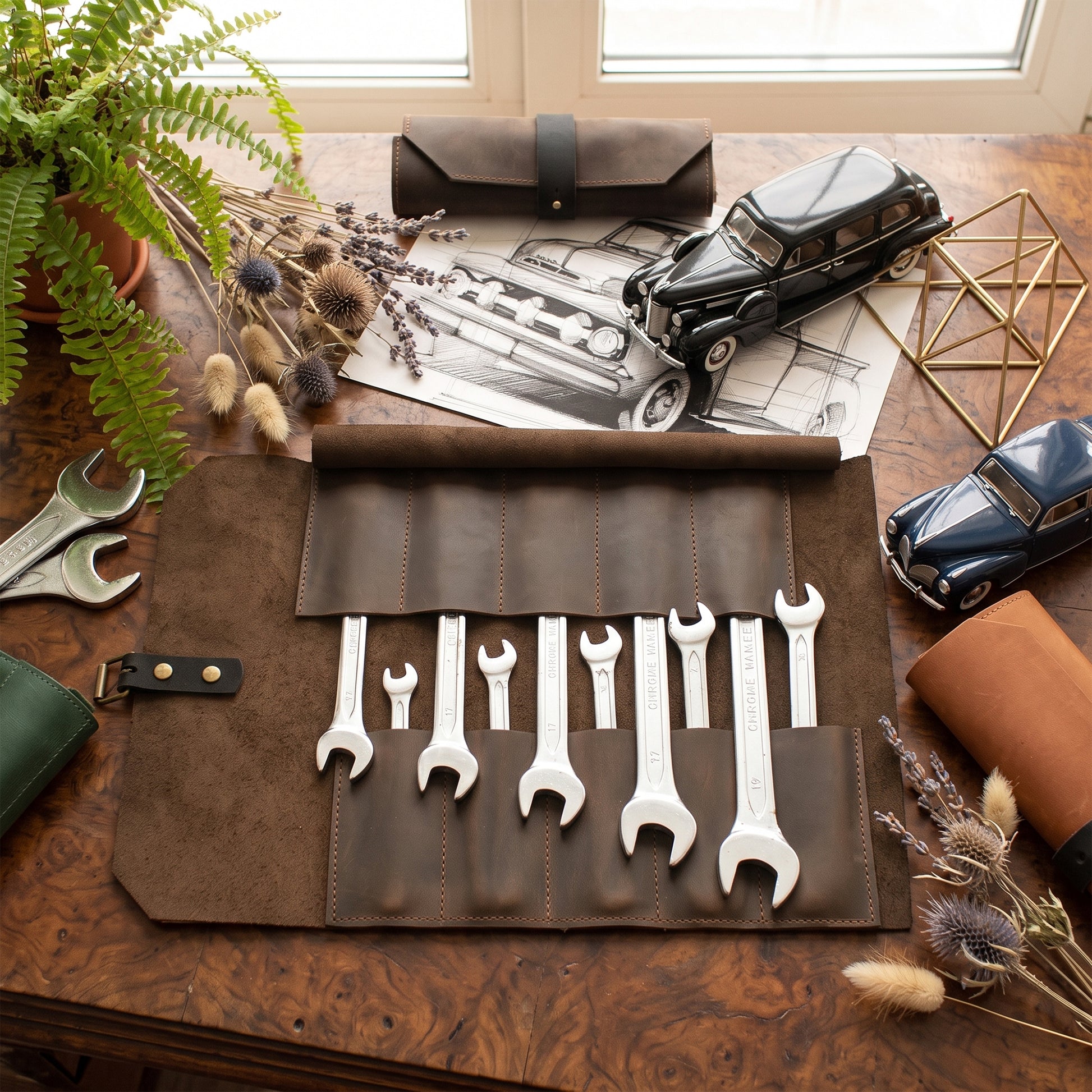A brown leather tool roll laid open on a wooden table, holding several silver wrenches. The scene includes a potted fern, dried flowers, toy cars, a black vintage car drawing, and metal wrenches as styled props.
