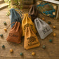 Five personalized drawstring pouches in different colors (blue, brown, yellow, gray, tan) arranged on a wooden tabletop with scattered polyhedral dice. The front center yellow pouch is engraved with 'Allistair' and a geometric emblem, and the front left tan pouch is engraved with 'Dino'.