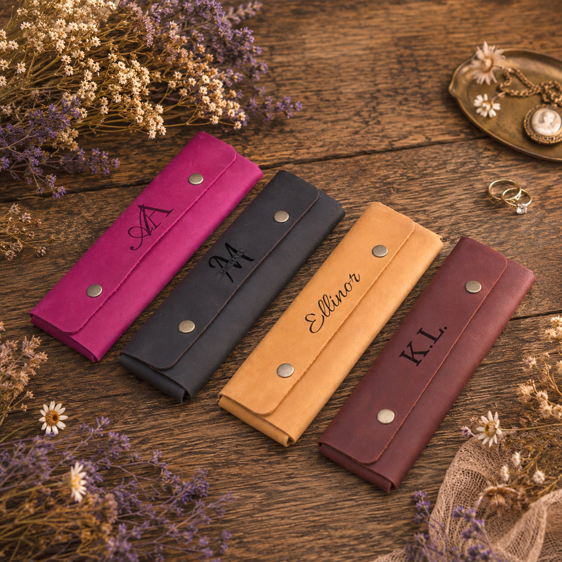 Four personalized leather wallet or card holder cases in pink, black, tan, and burgundy, arranged diagonally on a rustic wooden surface with dried flowers and jewelry props.