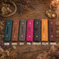 Six leather wallet or card holder cases in different colors are lined up on a rustic wooden surface with dried flowers and decorative props. The colors shown are denim blue, tan, gray, pink, black, yellow, and brown, each with a snap button closure and a personalized name or initial engraved on the front.