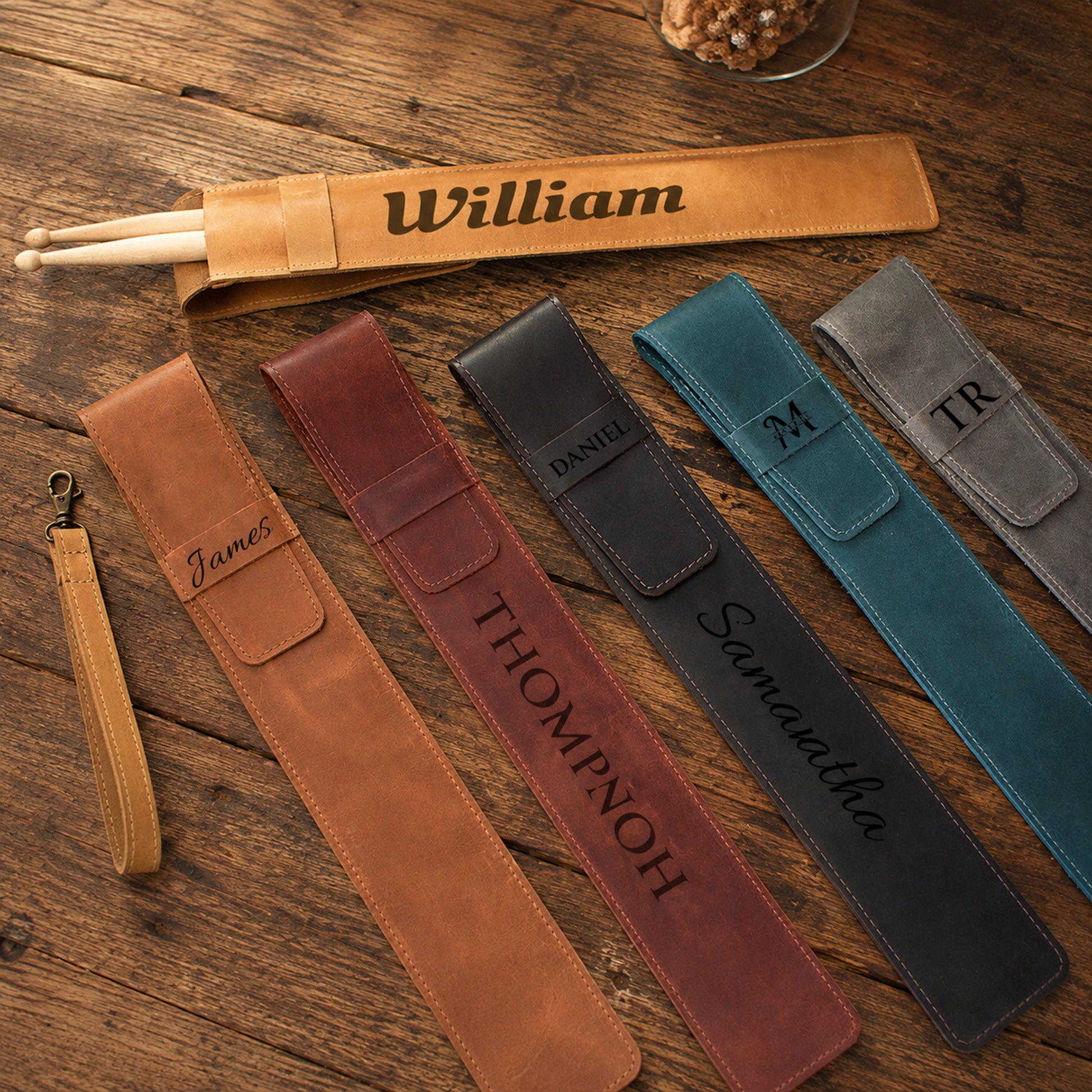 Five personalized leather drumstick pouches in different colors (tan, brown, black, teal, and gray) are arranged on a rustic wooden table. Each pouch features a custom engraved name (including William, James, Thompnoh, Daniel, and Samuelthka) and a matching wrist strap.