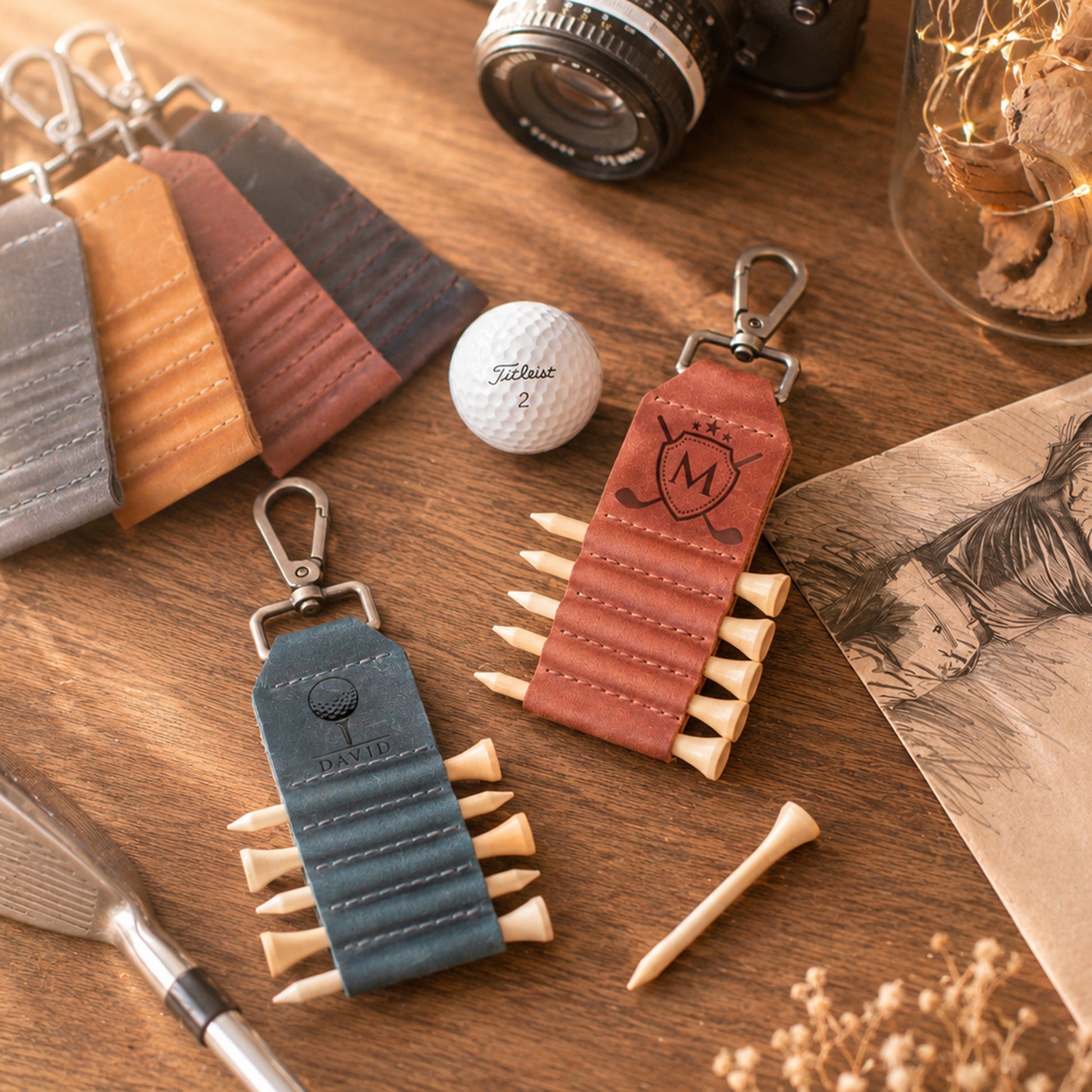 Two leather golf keychains styled on a wooden tabletop, one in slate blue and one in reddish-brown, both designed to look like rolled golf clubs with metal keyrings and faux tees. A golf ball, tees, camera, and other golf props are arranged around them.