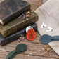 A styled flat-lay of leather keychain rings and matching card holders on a rustic wooden table, surrounded by vintage books, dried lavender, white wireless earbuds, and a beige linen cloth. One ring is orange with a circular 'RAG' logo, and the others are in dark green, slate blue, and dark brown.