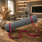 A gray textured yoga mat rolled up with a bright pink strap and wristband, displayed on a rustic wooden table in a warm, modern living room setting.