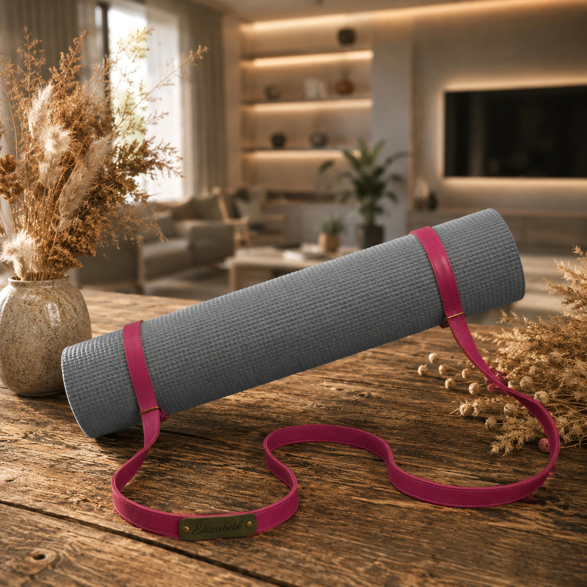A gray textured yoga mat rolled up with a bright pink strap and wristband, displayed on a rustic wooden table in a warm, modern living room setting.