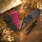 Four personalized leather pen sleeves in black, magenta, tan, and slate blue, arranged on a rustic wooden table with dried flowers, a book, and two black pens.