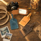 Three personalized leather luggage tags in blue, tan, and brown, styled on a wooden tabletop with a camera, rope, coins, polaroid photos, and a map.