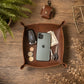 A brown leather square tray with raised edges, styled on a wooden tabletop with a smartphone, car key, coins, cash, keys, a small leather pouch, a fern, and dried flowers.