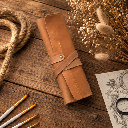 A rolled tan leather portfolio or scroll case with a wrap-around strap and metal snap, styled on a rustic wooden tabletop with dried flowers, rope, paintbrushes, and a sketchbook.