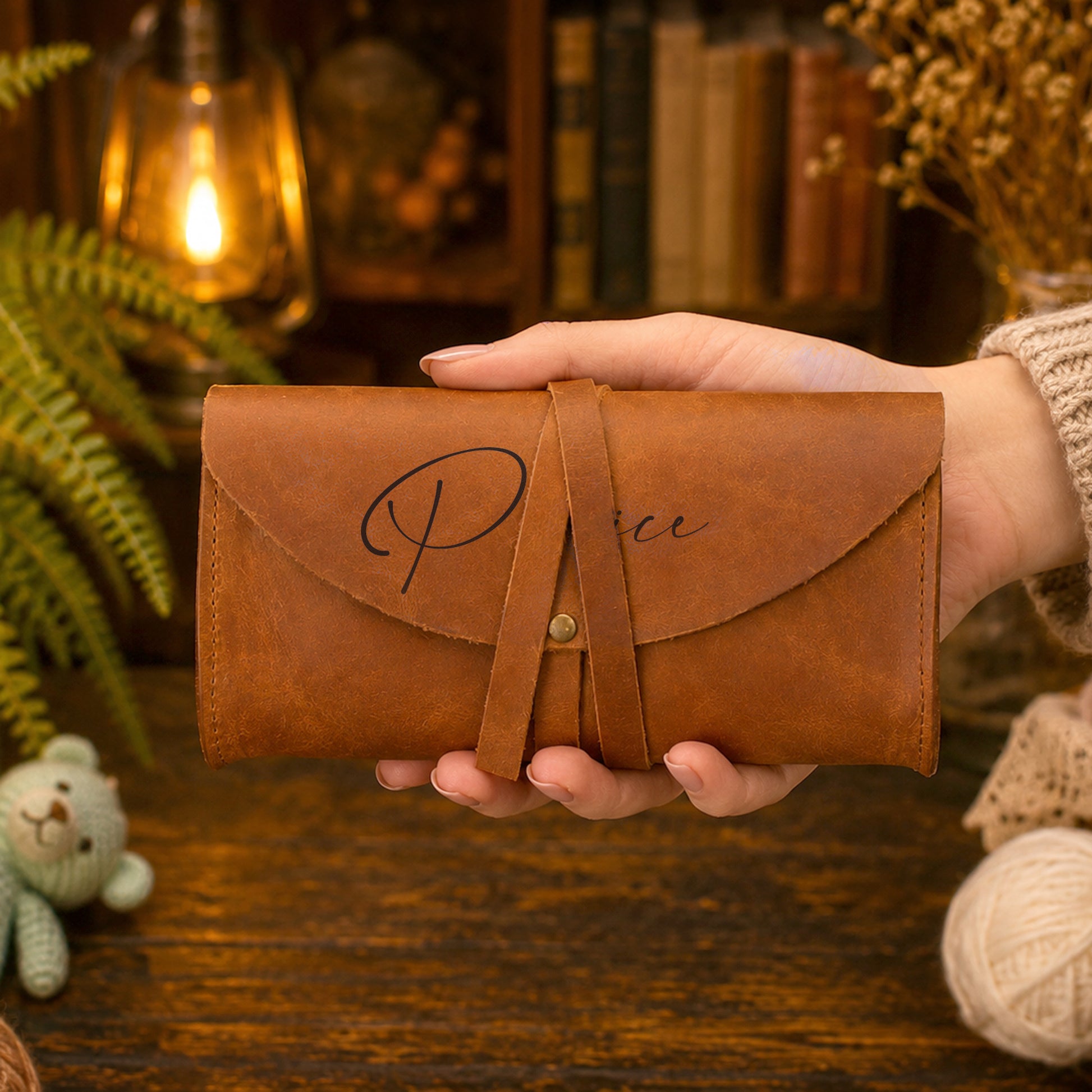 A hand holding a brown leather clutch wallet with a strap closure and cursive script on the front, set against a cozy, warm-toned background with books, a lamp, and dried flowers.