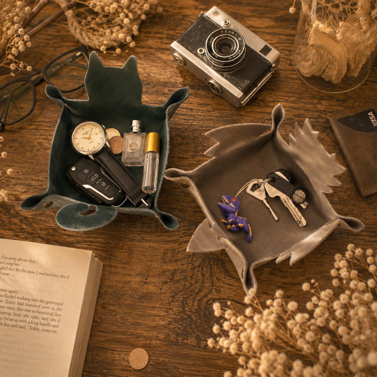 Two sculptural fox-shaped tray organizers styled on a wooden tabletop with vintage props including a camera, book, glasses, and dried flowers. One tray is dark teal and holds a watch, perfume bottle, keys, and wallet.