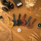 Six personalized leather keychains shaped like guitars are arranged in a row on a wooden tabletop, surrounded by vintage-style props including a camera, coins, a key, a compass, and dried wheat.