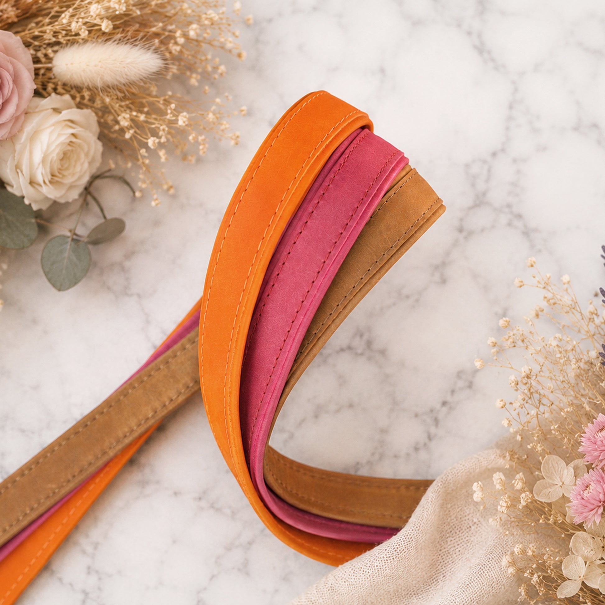 Three colorful belts (orange, pink, brown) on a marble surface with decorative flowers.