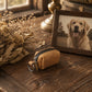 A small tan leather zip pouch with a metal key ring sits on a rustic wooden table beside a framed photo of a golden retriever, with dried flowers and warm indoor decor in the background.