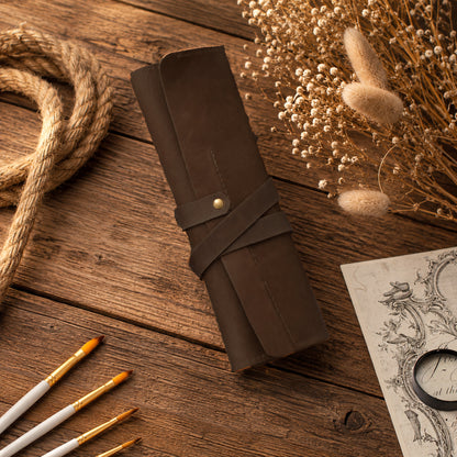 A dark brown leather scroll holder or canvas roll with a wrap-around strap and brass snap, displayed on a rustic wooden table with dried flowers, rope, paintbrushes, and a vintage-style sketchbook.