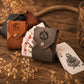 Two personalized card sleeves, one brown leather and one dark gray suede, displayed on a rustic wooden table with playing cards and dried flowers.