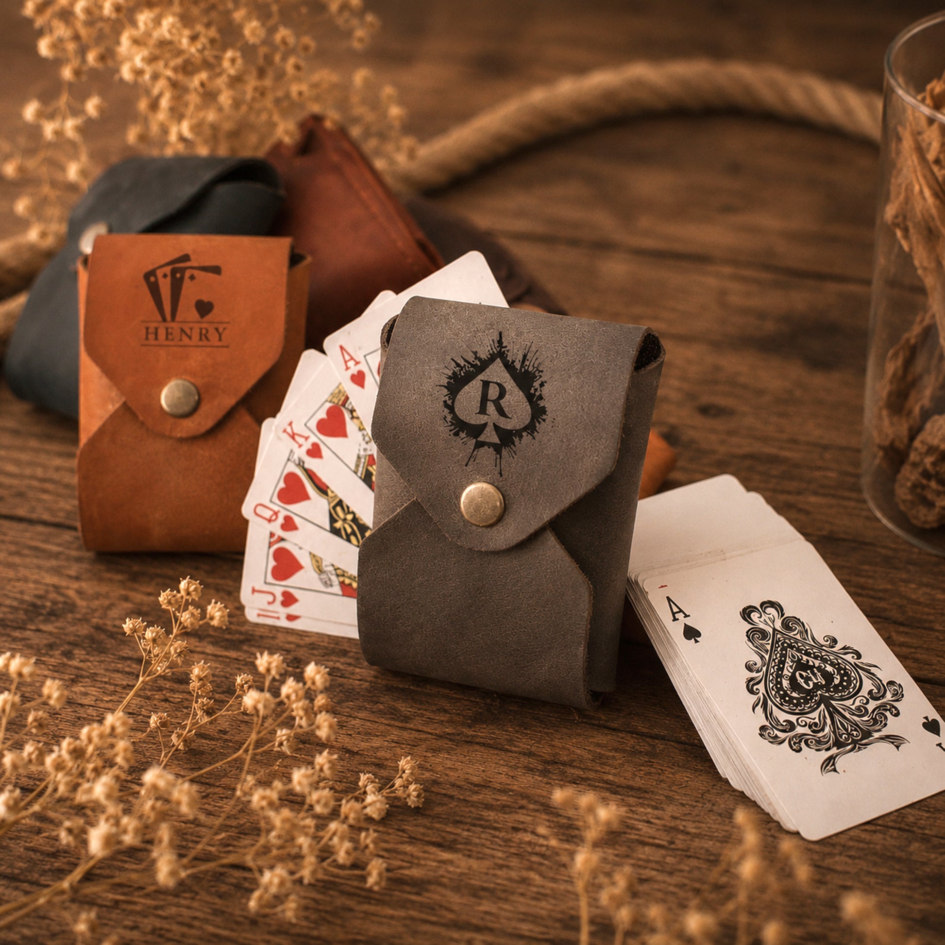 Two personalized card sleeves, one brown leather and one dark gray suede, displayed on a rustic wooden table with playing cards and dried flowers.