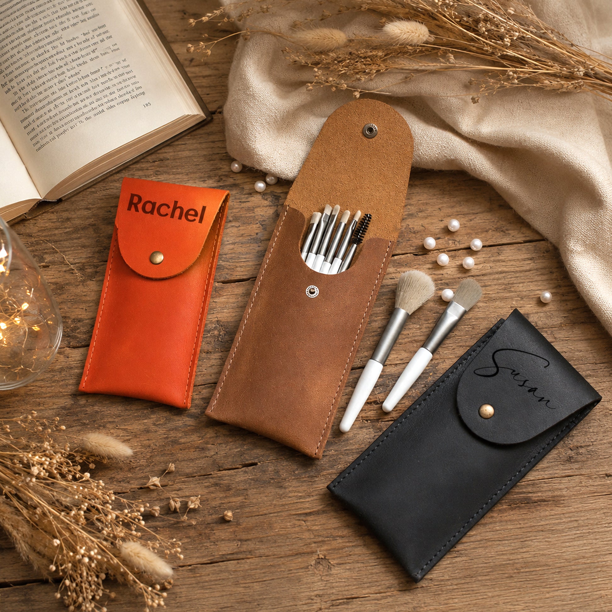 Three personalized leather makeup pouches in orange, brown, and black are arranged on a rustic wooden table with dried flowers, a book, and makeup brushes. The orange pouch is labeled 'Rachel' and the black pouch is labeled 'Susan'.