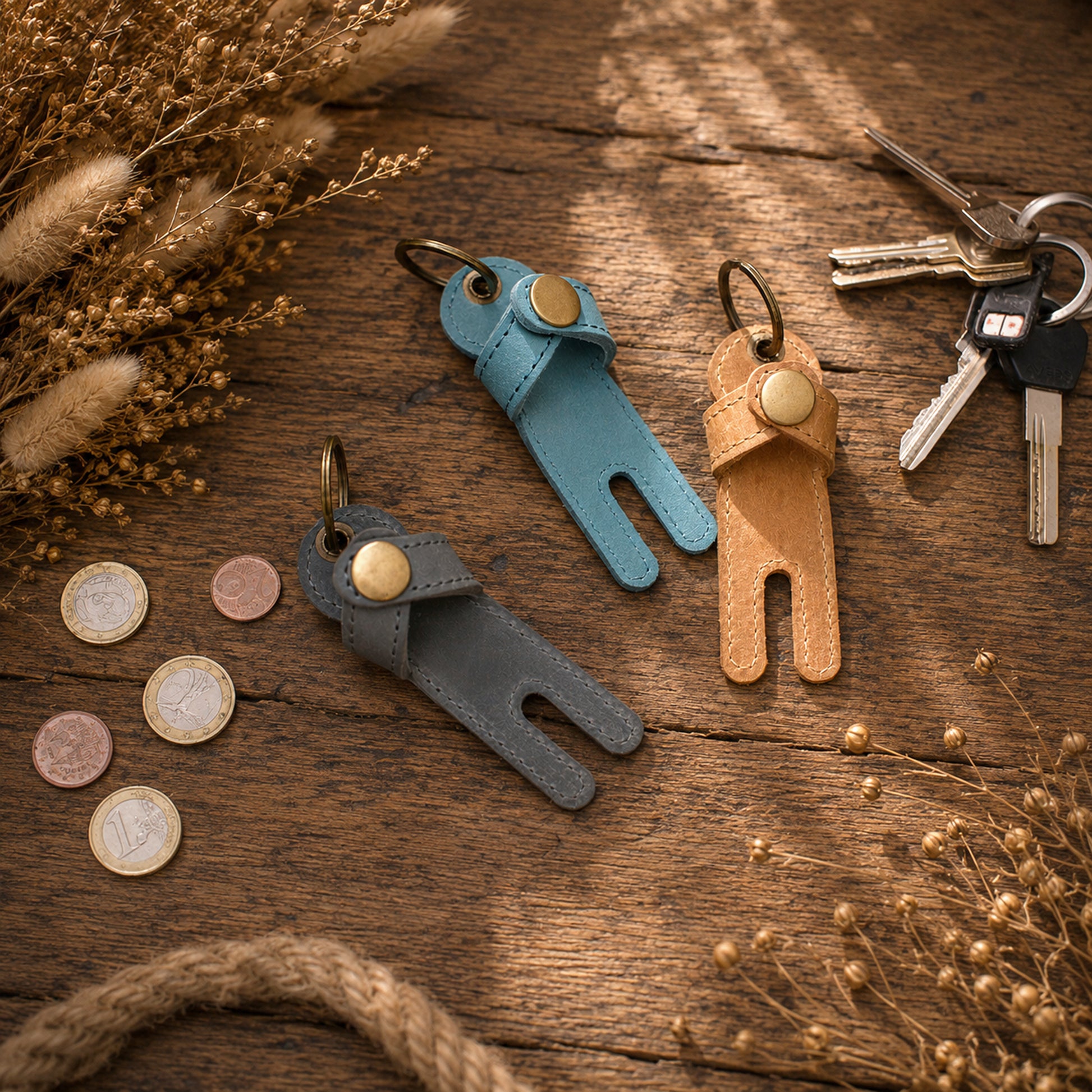 Three small leather keychain golf tees in blue, tan, and gray are arranged on a rustic wooden surface with dried grasses, coins, rope, and a set of keys nearby.