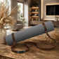 A gray textured yoga mat with brown straps and a silver metal plate engraved with 'YOGA', resting on a rustic wooden table in a warm, modern living room setting.