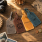Four personalized leather luggage tags in gray, brown, mustard yellow, and teal, arranged on a rustic wooden table with travel-themed props like a camera, rope, and coins.