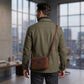 Man wearing a green jacket with a brown leather bag in an indoor setting with large windows.