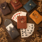 Six personalized card sleeves in different colors (brown, blue, orange, gray, and mustard yellow) are arranged on a rustic wooden table with playing cards and dried flowers. Some sleeves are personalized with names like 'Frannie', 'HARRY', and 'HENRY', and feature brass snap buttons.