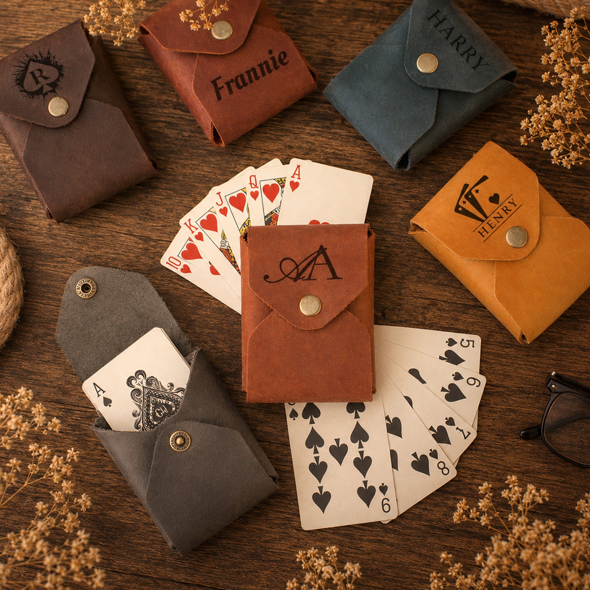 Six personalized card sleeves in different colors (brown, blue, orange, gray, and mustard yellow) are arranged on a rustic wooden table with playing cards and dried flowers. Some sleeves are personalized with names like 'Frannie', 'HARRY', and 'HENRY', and feature brass snap buttons.
