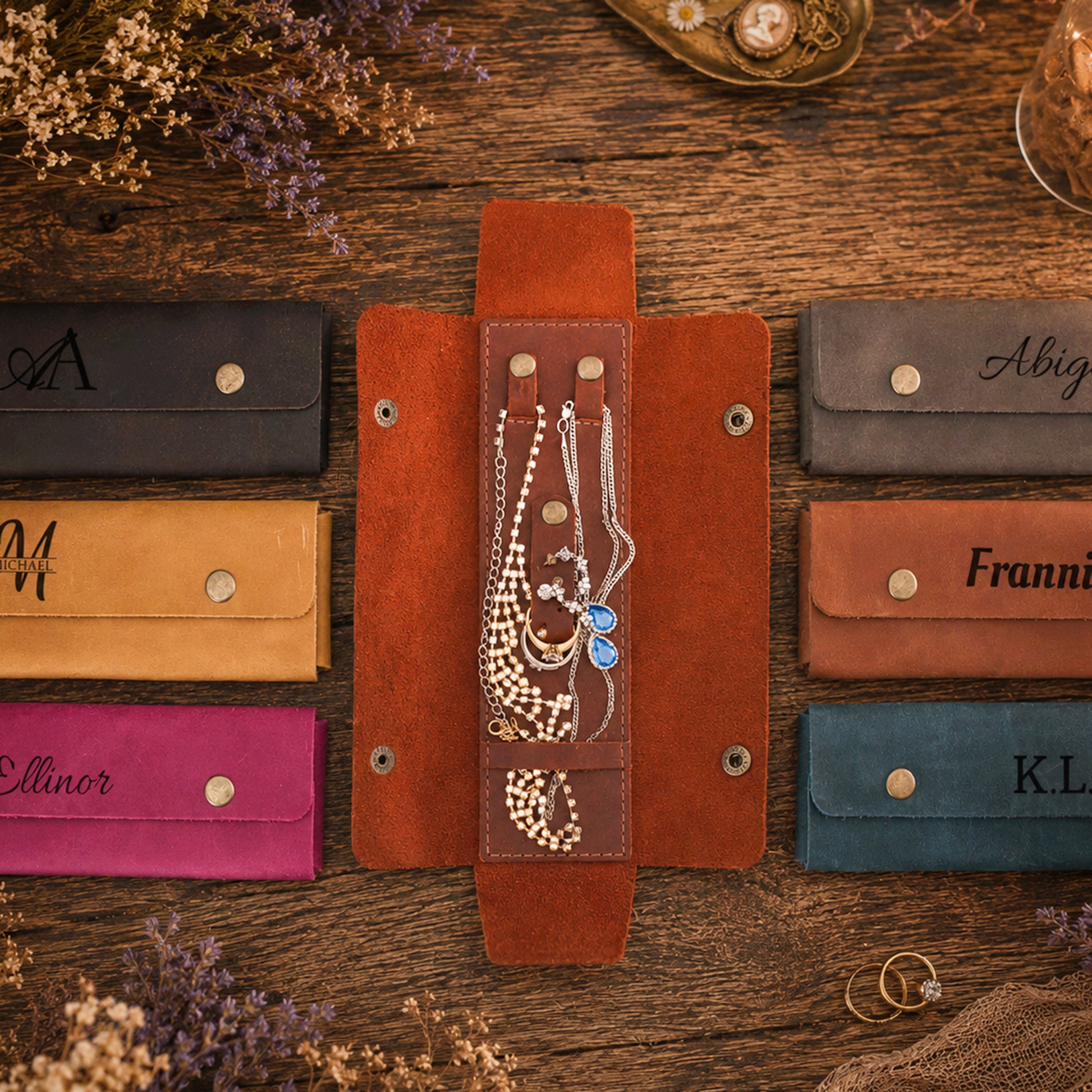 A set of personalized leather jewelry organizer sleeves displayed on a rustic wooden table. The center sleeve is open, showing a brown leather interior with a vertical ring holder containing necklaces and bracelets, flanked by six closed sleeves in different colors (black, tan, brown, pink, gray, rust, and teal) each personalized with an elegant script name.