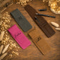 Four personalized leather pen cases in dark gray, brown, tan, and magenta pink, arranged on a rustic wooden table with dried flowers and pens.