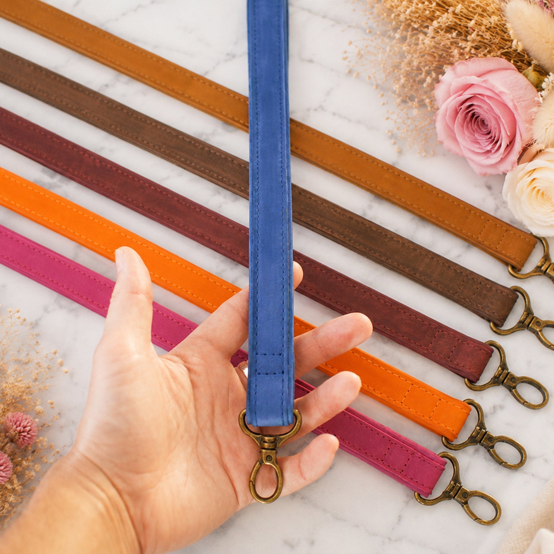 Collection of colorful leather straps with a hand holding a blue strap on a marble surface with flowers.