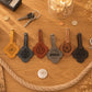 Six personalized leather key tags arranged on a wooden tabletop, each with a different monogram or name (AJF, M&G, D, William, W, and Abigail). The tags are shown in various colors including mustard yellow, dark gray, reddish-brown, silver, tan, and black, with keys and decorative props around them.