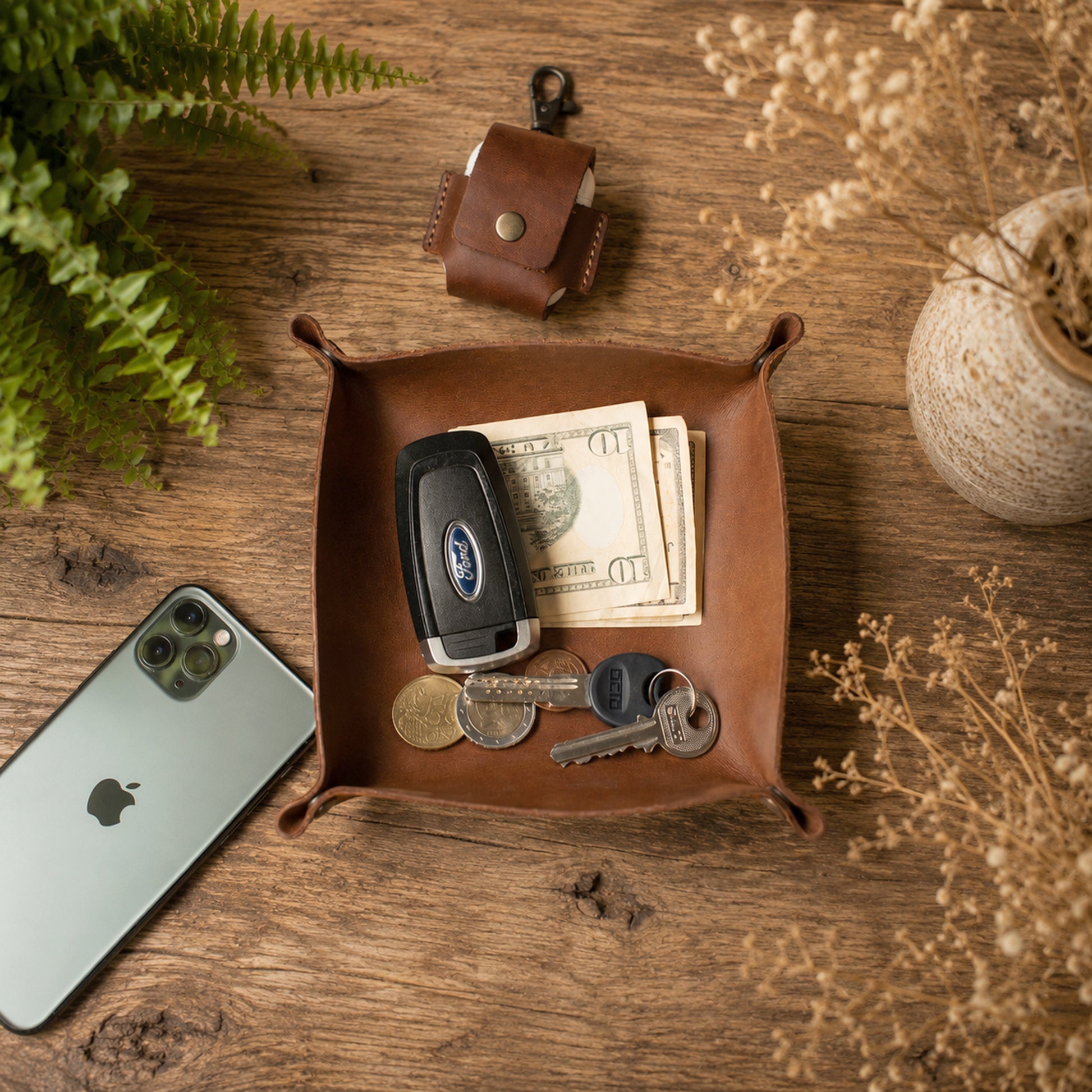 A brown leather square tray with rolled corners and side loops, styled on a rustic wooden table. The tray contains a car key fob, cash, and keys, with a small matching leather pouch above it, a smartphone to the left, and dried flowers in a vase to the right.