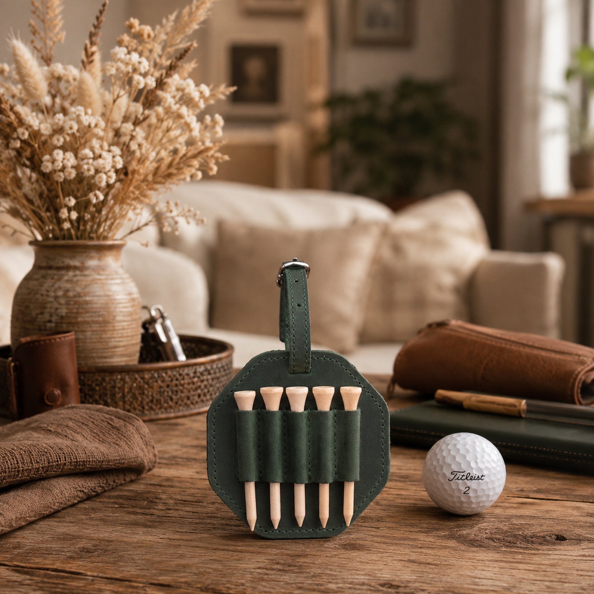 A dark green leather golf club addresser set with wooden tees, displayed on a rustic wooden table next to a golf ball, with a cozy living room background.