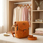 Orange handbag with 'AGH' logo on a bed in a room with clothes and shelves.