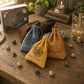 Three personalized drawstring pouches in brown, blue, and yellow leather-like material with printed names and crest designs, arranged on a rustic wooden table with dice and books in the background.