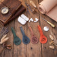Four personalized leather luggage tags in green, blue, brown, and orange, arranged on a rustic wooden table with styled flat-lay props including a leather journal, earbuds, a rolled tan leather sheet, and dried flowers.