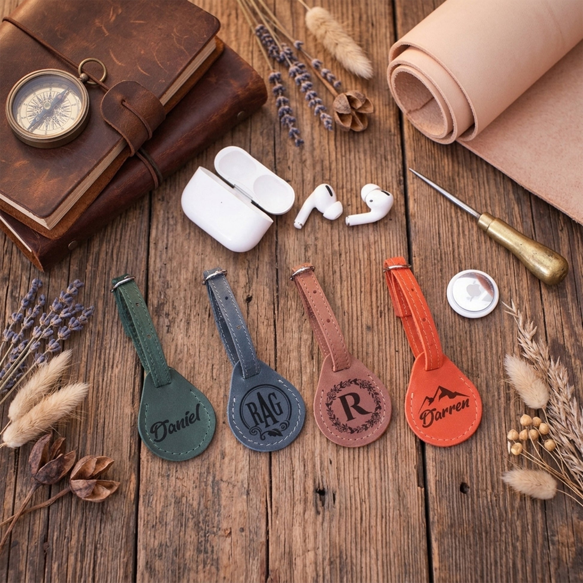 Four personalized leather luggage tags in green, blue, brown, and orange, arranged on a rustic wooden table with styled flat-lay props including a leather journal, earbuds, a rolled tan leather sheet, and dried flowers.