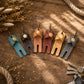 Five leather keychain pouches in assorted colors (teal, charcoal, tan, burgundy, and gray) arranged on a rustic wooden surface with dried flowers and keys as props.