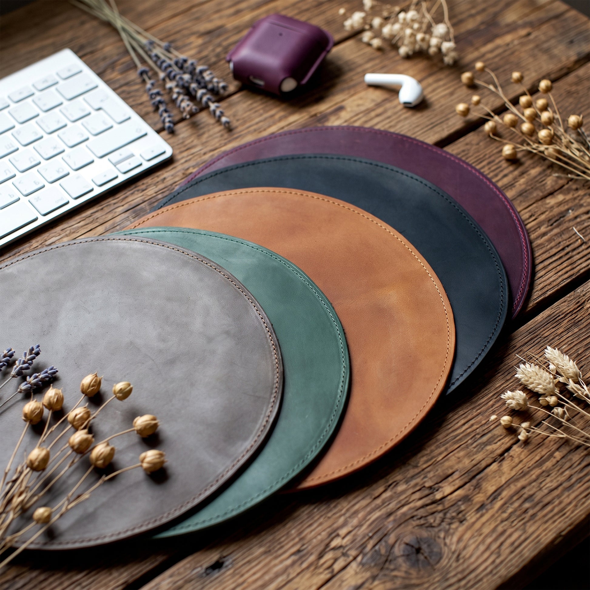 Four round leather desk mats in gray, green, tan, and burgundy, arranged in a fan shape on a rustic wooden table with dried flowers, a keyboard, and earbud case as props.
