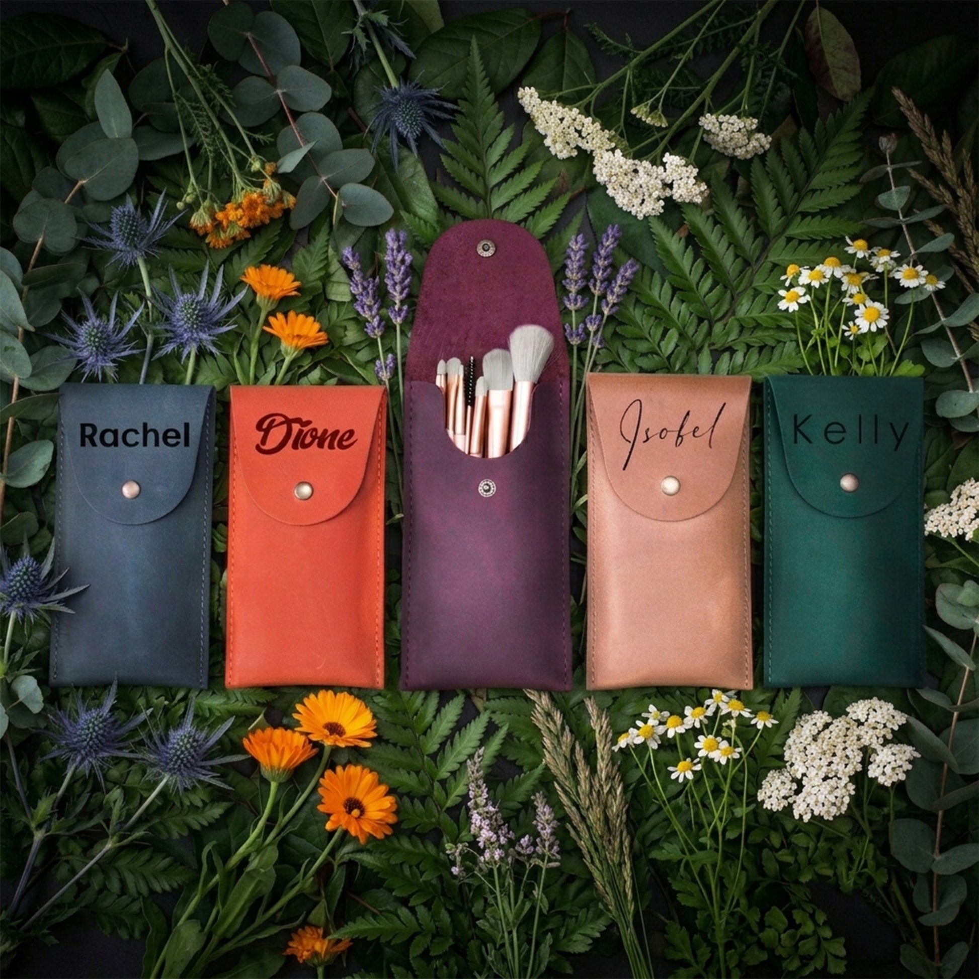 Four personalized makeup brush pouches in dark blue, orange, plum purple, and forest green, arranged in a flat-lay with makeup brushes in the center pouch, surrounded by green foliage and small wildflowers. The pouches are personalized with the names Rachel, Diane, Isobel, and Kelly.