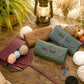 Four leather crochet hook storage cases in brown, green, and blue-gray, each personalized with a name (Kelly, Patience, Rachel) and styled on a rustic wooden table with yarn, knitting needles, a lantern, and greenery.