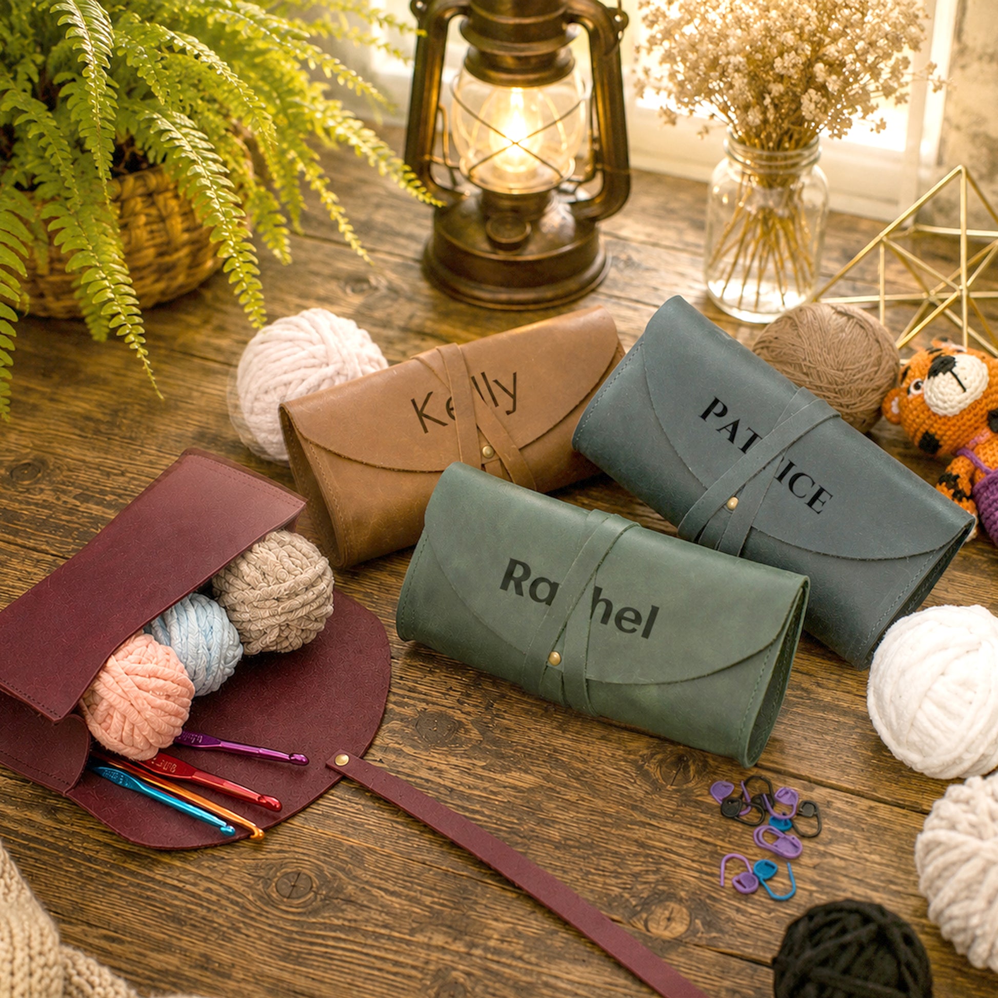 Four leather crochet hook storage cases in brown, green, and blue-gray, each personalized with a name (Kelly, Patience, Rachel) and styled on a rustic wooden table with yarn, knitting needles, a lantern, and greenery.