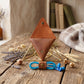 A brown leather geometric phone pouch with a gold metal frame stand, shown on a rustic wooden table with dried flowers, wheat, and a blue cord accessory.
