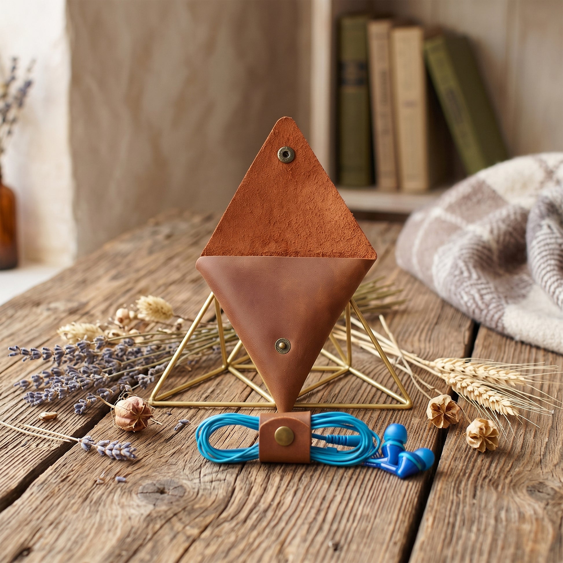 A brown leather geometric phone pouch with a gold metal frame stand, shown on a rustic wooden table with dried flowers, wheat, and a blue cord accessory.