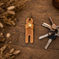 A tan leather keychain holder shaped like a small figure with a gold snap button, displayed on a rustic wooden surface next to a set of keys and dried decorative grass.