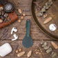 A dark green leather keychain or bag charm with a loop strap, styled in a flat-lay arrangement on a rustic wooden table with various decorative props including a leather case, AirPods, a compass, and dried botanicals.